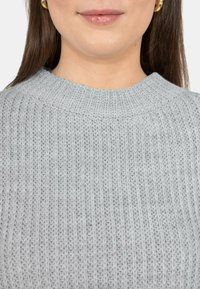 Primo piano della parte inferiore del viso e del collo di una donna che indossa orecchini a cerchio d'oro e un maglione grigio chiaro lavorato a maglia.