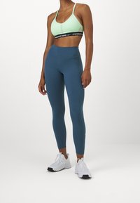Donna che indossa un reggiseno sportivo verde menta, leggings blu a vita alta e sneaker atletiche bianche su uno sfondo bianco.
