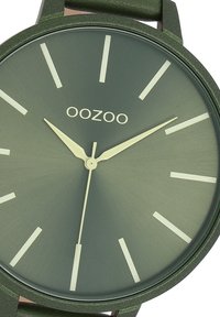 Montre analogique verte avec un fini mat, des repères horaires blancs et des aiguilles fines. Le nom de la marque "OOZOO" est affiché sur le cadran. Bracelet en cuir.