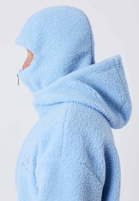 Personne portant une veste polaire bleu clair avec capuche attachée couvrant la tête et une partie du visage, montrée de profil sur fond blanc.
