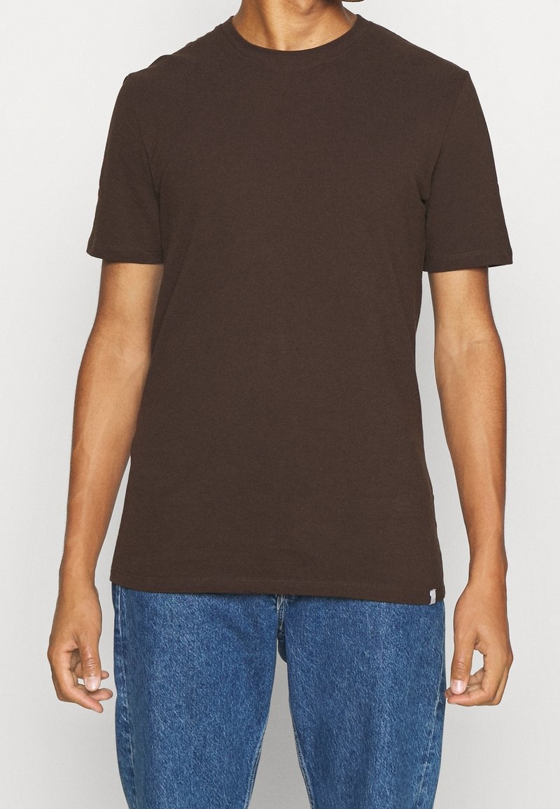 T-shirt en coton marron foncé avec des manches courtes et un col rond, associé à un jean en denim bleu. Design simple en couleur unie, sans motifs visibles.