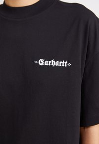 Svart bomulls t-shirt med korta ärmar, med en vit "Carhartt"-logotyp i en stiliserad font på vänster bröstområde. Slät textur.
