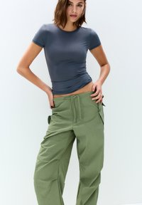 Siva prilegajoča majica s kratkimi rokavi v kombinaciji z ohlapnimi olivno zelenimi cargo hlačami, ki imajo vrvico za zategovanje in stranske žepe. Gladka tekstura.