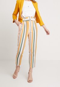 Blazer giallo sopra una maglia bianca abbinato a pantaloni a vita alta a righe gialle, verdi e bianche. Presenta una cintura e un design corto.
