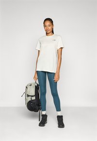 Jasny T-shirt z małym logo, turkusowe legginsy, czarne buty trekkingowe i szary plecak z czarnymi akcentami. Prosty design i funkcjonalne cechy.