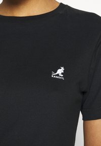 Sort bomulds-T-shirt med rund hals, der har et lille hvidt kænguru-logo på venstre bryst. Glat tekstur og korte ærmer.