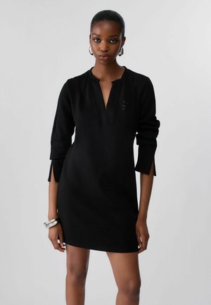 Giovane donna che indossa un vestito nero al ginocchio con maniche a tre quarti, orecchini a cerchio argentati e braccialetti argentati a un polso.