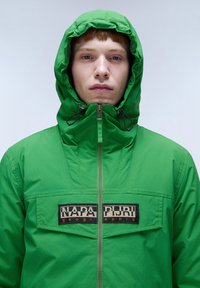 Grøn hættejakke med lynlås foran, prydet med et stort sort og beige logo patch. Glat, letvægtsstof og justerbare snore i hætten.