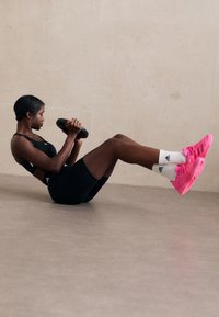 Atleta che esegue un esercizio su una superficie, indossando un top corto nero e pantaloni corti, con scarpe rosa e calze bianche Adidas, mentre tiene un peso nero.
