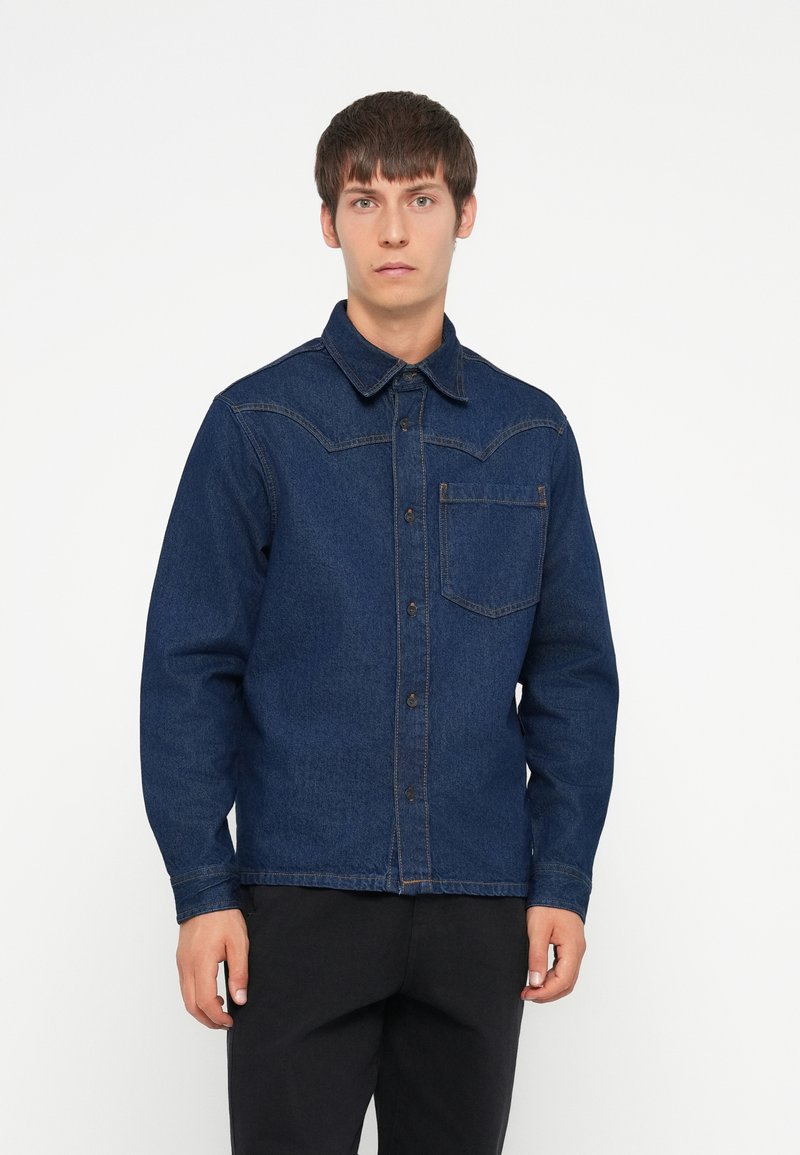 Giacca in denim blu scuro con chiusura a bottoni, colletto aperto e una tasca sul petto. Presenta cuciture a contrasto e una vestibilità comoda.
