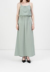 Robe maxi verte avec un motif à pois, ceinture élastique, bretelles spaghetti et une silhouette fluide. Assortie à des chaussures noires à bout pointu.