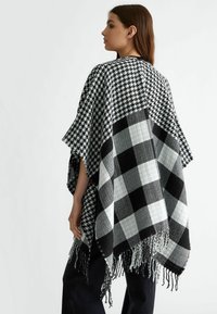 Poncho a righe in bianco e nero con design a scacchi houndstooth e motivo a quadretti, con bordi frangiati e apertura frontale.