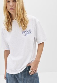 T-shirt blanc à manches courtes avec une coupe décontractée et un texte brodé bleu "Game of Hearts". Tissu doux, col rond, design minimal.