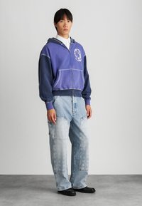 Hettejakke med glidelås i lilla og blåtoner med et astronautmotiv. Kombinert med lyseblå, baggy jeans med lommer. Svarte sko.