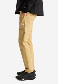 Pantaloni cargo beige con multiple tasche, realizzati in tessuto leggero. Indossati con scarpe da trail nere e testurizzate e una giacca nera.