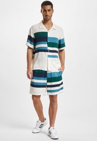 Lyhythihainen nappipaita ja shortsit, joissa on vaakaraitoja väriltään teal, laivastonsininen ja valkoinen. Kangas näyttää kevyeltä ja sileältä.