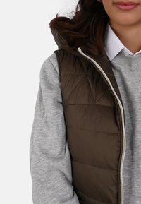 Woman wearing a brown quilted vest over a grey sweater and white collared shirt, showing upper torso and part of the face.