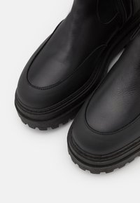 Mustat nahkakengät paksuilla pohjilla, pyöreillä varpailla ja ommelluilla yksityiskohdilla sileillä yläpintoilla.