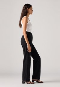 Zwarte wijde jeans met hoge taille, gemaakt van denim, gecombineerd met een witte aansluitende tanktop en zwarte sandalen met hakken en bandjes.