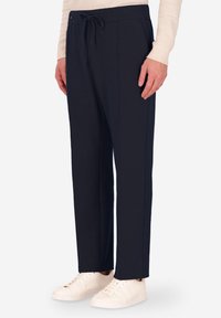 Pantaloni tuta in navy con cintura elastica e cordoncino regolabile. Presentano tasche laterali e una vestibilità slim. Abbinati a sneakers chiare.