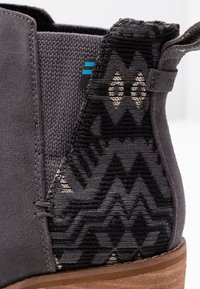 Botín de ante gris con un panel textil negro texturizado y estampado, con una suela de madera marrón y un sutil acento azul.