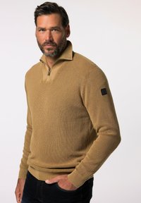 Beige gestrickter Pullover mit einem Halbreißverschluss-Kragen, der eine strukturierte Stoffqualität und ein schwarzes Logo-Patch an der linken Ärmeln aufweist.