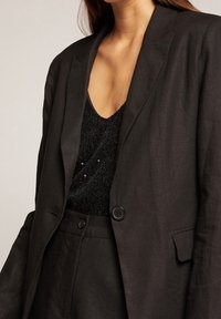 Černý lněný blazer s klopou, zapínání na jeden knoflík a bočními kapsami, nošený přes černý top se sequiny, zdůrazňující štíhlou siluetu.