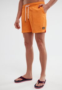 Oranžinės maudymosi šortai su elastingu juosmeniu ir balta raišteliu. Su šoninėmis kišenėmis ir mažyte juoda logotipo etikete. Dominuoja tamsiai mėlyni šlepetės.