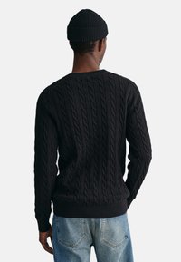 Schwarzer Kabelstrickpullover mit strukturierten Mustern, gerippten Bündchen und Saum, kombiniert mit hellblauen Jeans und einer passenden schwarzen Mütze.
