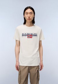 Ung mand med glat sort hår iført en hvid Napapijri T-shirt med logo og khaki cargo-bukser foran en ensfarvet lys baggrund.