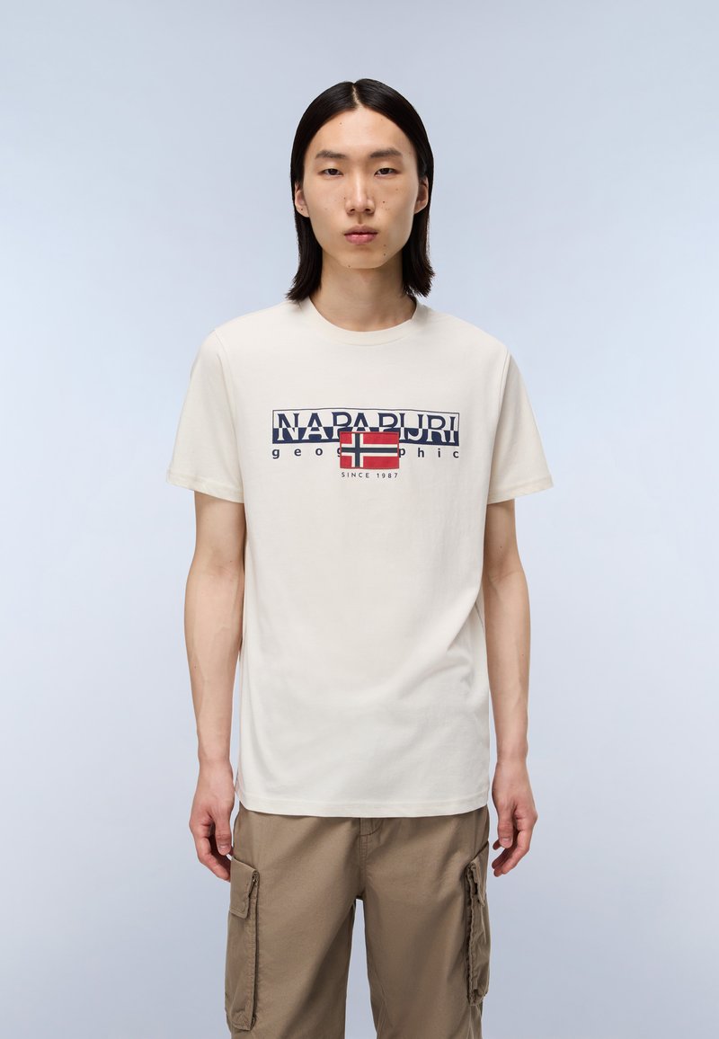 Ung mand med glat sort hår iført en hvid Napapijri T-shirt med logo og khaki cargo-bukser foran en ensfarvet lys baggrund.
