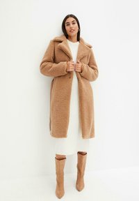 Cappotto teddy marrone con texture, colletto a punta, bottoni frontali e tasche laterali, indossato sopra un vestito bianco e abbinato a stivali beige fino al ginocchio.