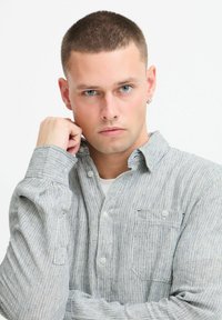 Striped button-up shirt in gray and white fabric, featuring a chest pocket and rolled sleeves. Model has short hair and an earring.
