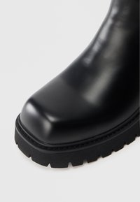 Musta nahkainen saapas, jossa on pyöreä kärki, mattapintainen viimeistely ja teksturoitu kumipohja näkyvällä kuvioinnilla. Ompeleet pohjassa koristeena.