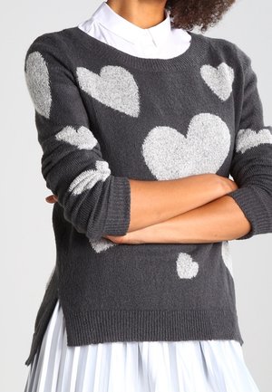 Dunkelgrauer gestrickter Pullover mit großen weißen Herzmustern, getragen über einem weißen Hemd mit Kragen und Rock, Arme vor der Brust verschränkt.