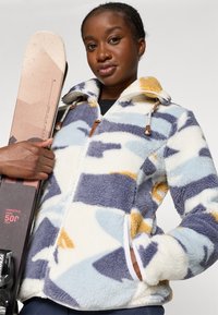 Fleece jack in crème, blauw en geel abstract patroon; heeft een ritssluitingen, zakken en decoratieve leren accenten. Model houdt ski's vast.
