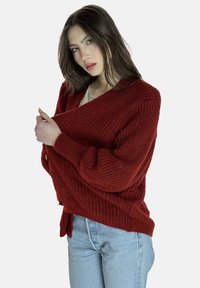 Gebreid vest in roestkleur met een gestructureerd patroon, oversized mouwen en een V-hals ontwerp. Knoopdetail aan de voorkant.