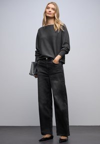 Šedý, oversize svetr s dlouhými rukávy, zkombinovaný s černými džínami s vysokým pasem a širokým střihem. Černé baleríny, držící stříbrnou kabelku.