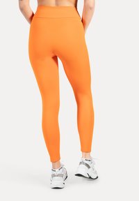 Heldere oranje legging met een gladde, rekbare textuur en een hoge tailleband, gecombineerd met witte en grijze sportschoenen.