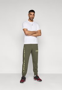 Λευκό βαμβακερό t-shirt με μικρό μαύρο λογότυπο, χακί παντελόνι με κίτρινο κείμενο «Jordan» και μαύρα/κόκκινα αθλητικά παπούτσια με υφασμάτινες λεπτομέρειες.