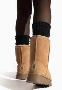Botas de ante marrón hasta la rodilla con un puño de punto negro texturizado, que presentan un detalle de logo en el costado y una suela gruesa y resistente.