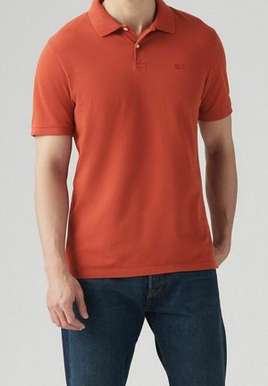 Homme portant un polo orange brûlé à manches courtes et un jean bleu foncé, debout devant un fond gris clair uni.