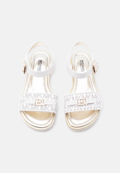 Λευκά σανδάλια Michael Kors με λουράκια στον αστράγαλο, πλατιές μπροστινές λωρίδες με σχέδιο λογότυπου MK και χρυσό έμβλημα MK σε κάθε σανδάλι.