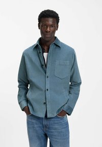 Hellblaues Cordhemd mit einer Brusttasche, schwarzen Knöpfen und lockerer Passform, getragen über einem weißen Unterhemd und kombiniert mit Jeans.