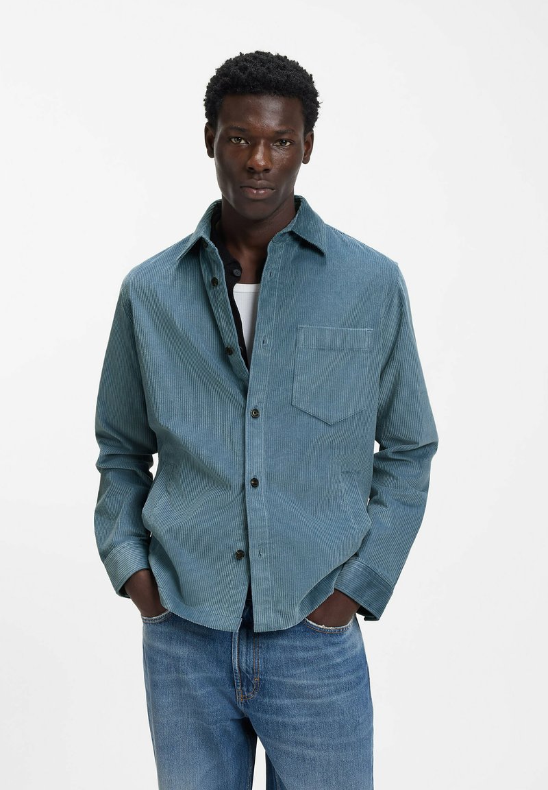 Hellblaues Cordhemd mit einer Brusttasche, schwarzen Knöpfen und lockerer Passform, getragen über einem weißen Unterhemd und kombiniert mit Jeans.