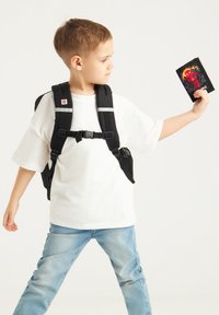 Czarny plecak z odblaskowymi paskami noszony przez dziecko w białym T-shircie i jasnoniebieskich dżinsach, trzymające dekoracyjną kartkę z żywymi grafikami.