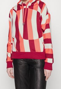 Sudadera con capucha y rayas llamativas en rojo, naranja y crema. Cuenta con capucha con cordón, puños acanalados y un corte relajado.