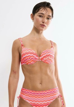 Woman wearing pink, orange, and white zigzag patterned bikini, standing against a plain white background.