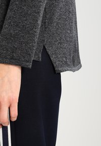 Graues, strukturiertes Langarmoberteil mit seitlichen Schlitzen, kombiniert mit marineblauen Hosen, die an den Seiten weiße Streifen aufweisen.