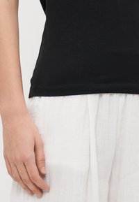 T-shirt de algodão preto com corte reto e decote redondo, combinado com calças largas de tecido texturizado brancas. Close-up de uma mão repousando sobre o tecido.